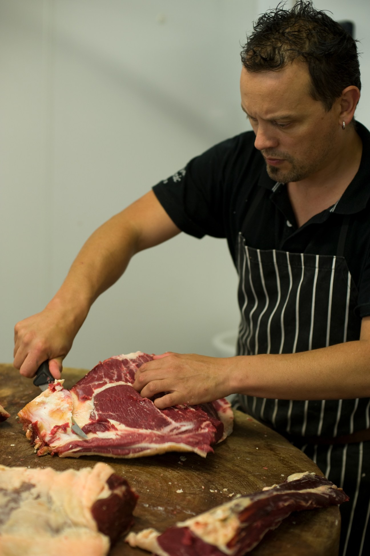 A butcher in a striped apron cuts raw meat on a wooden board.