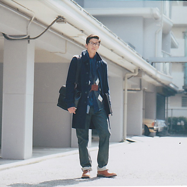 Man in dark Sacai coat and cardigan with Hermès scarf and Comme des Garçons shoes standing in modern interior