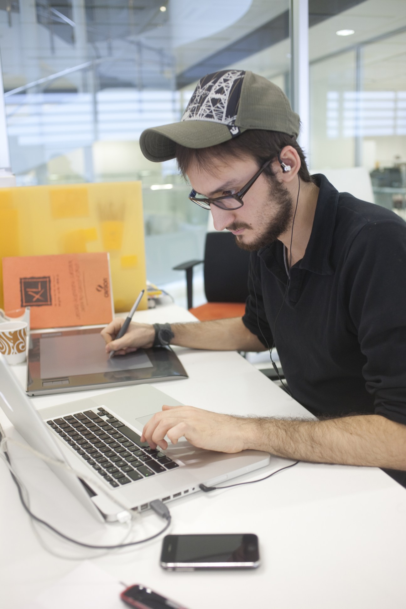 A man wearing glasses and a cap works at a laptop in a modern office with colorful design elements visible in the background.