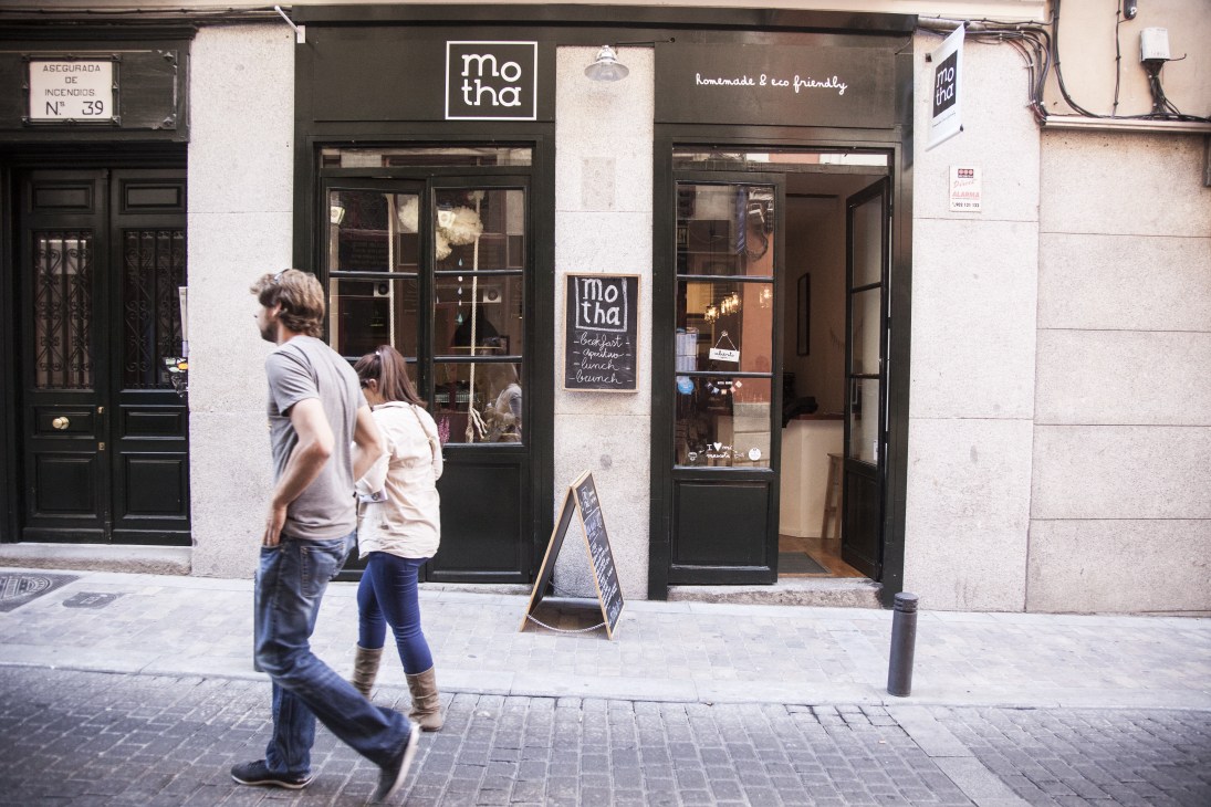 Pedestrians walk past Motha café storefront with black windows and eco-friendly signage in Madrid.
