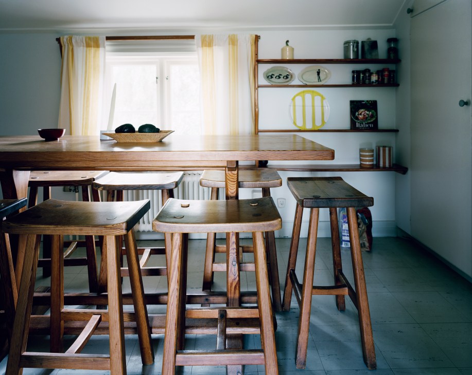 Minimalist dorm room with wooden table, chairs and stools in natural tones, white walls, shelving and yellow striped curtains.