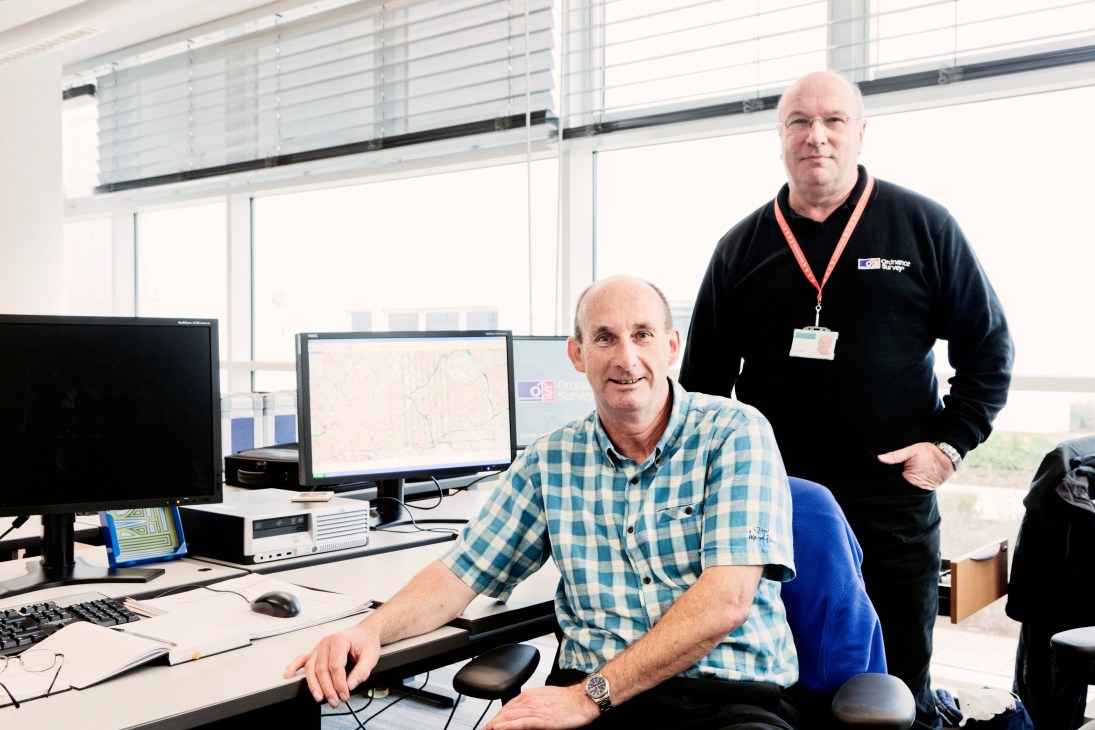 Two Ordnance Survey staff members, John McGrandle and Ron Cole, pose in an office with computer workstations displaying maps.
