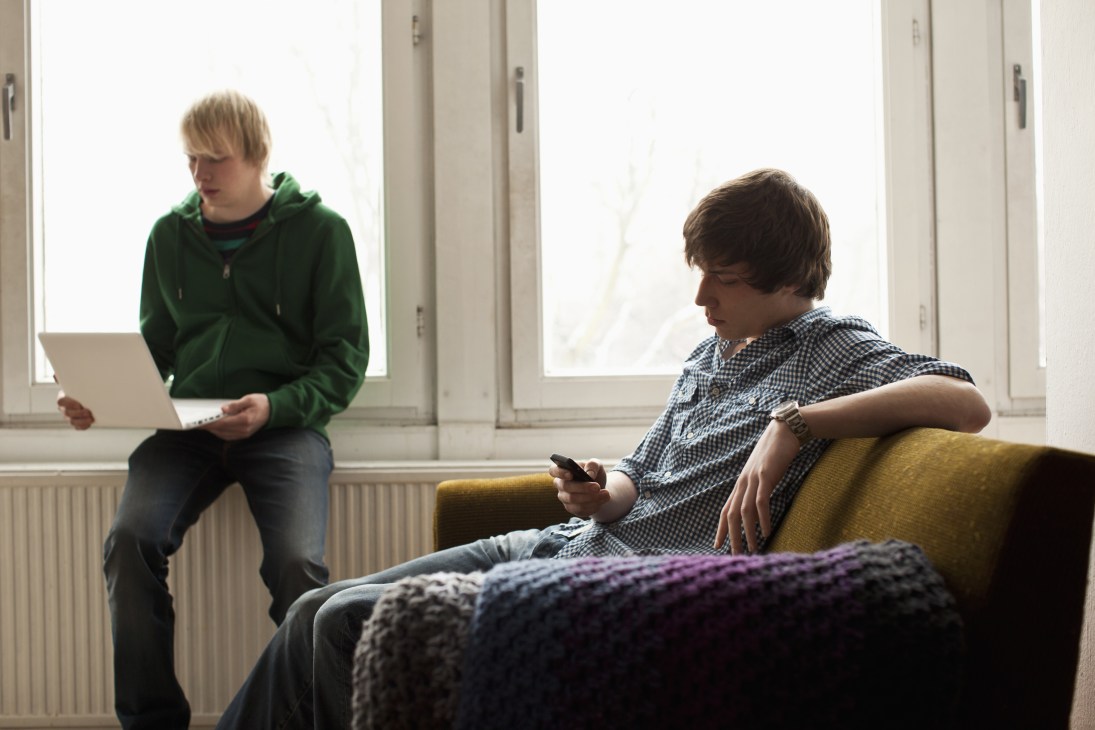 Two young men in a bright room, one holding a laptop and one holding a smartphone on a couch.