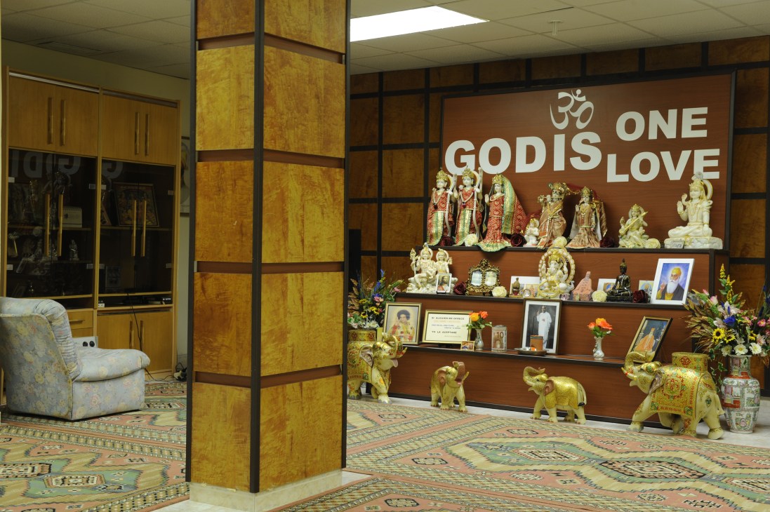 Interior of Hindu temple altar with golden deity statues and 'One God Is One Love' text on brown wall.