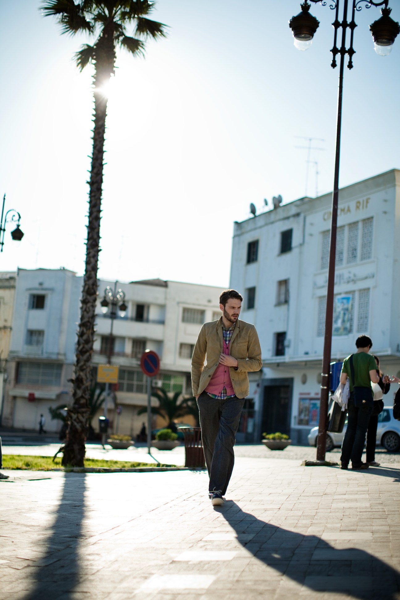 Man wearing a tan jacket and cardigan walking through a sunny Tangier square with palm trees and colonial-era buildings.
