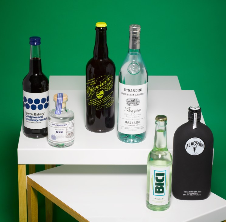 Bottles and glasses arranged on white surfaces against a green background for a Nordic Mojito cocktail recipe.