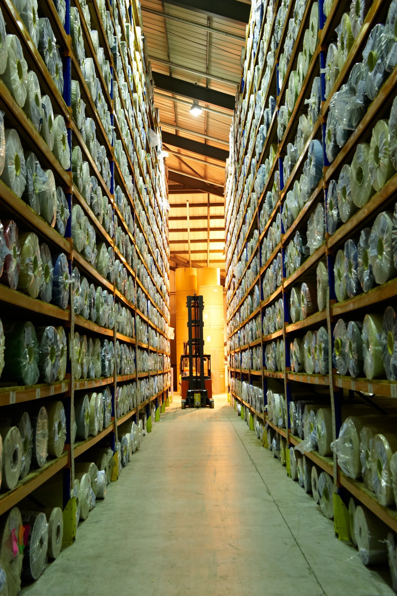 Warehouse aisle lined with thousands of colored paper rolls stacked on yellow shelves, with a forklift visible in the distance.