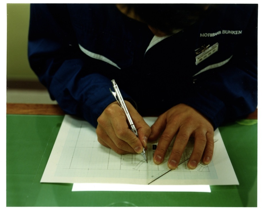 Designer's hands drawing kanji characters on graph paper with mechanical pencil and ruler.