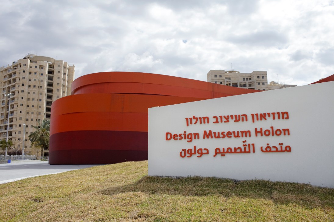 Design Museum Holon's distinctive red curved metal structure with signage in Hebrew, English, and Arabic against residential buildings in Israel.