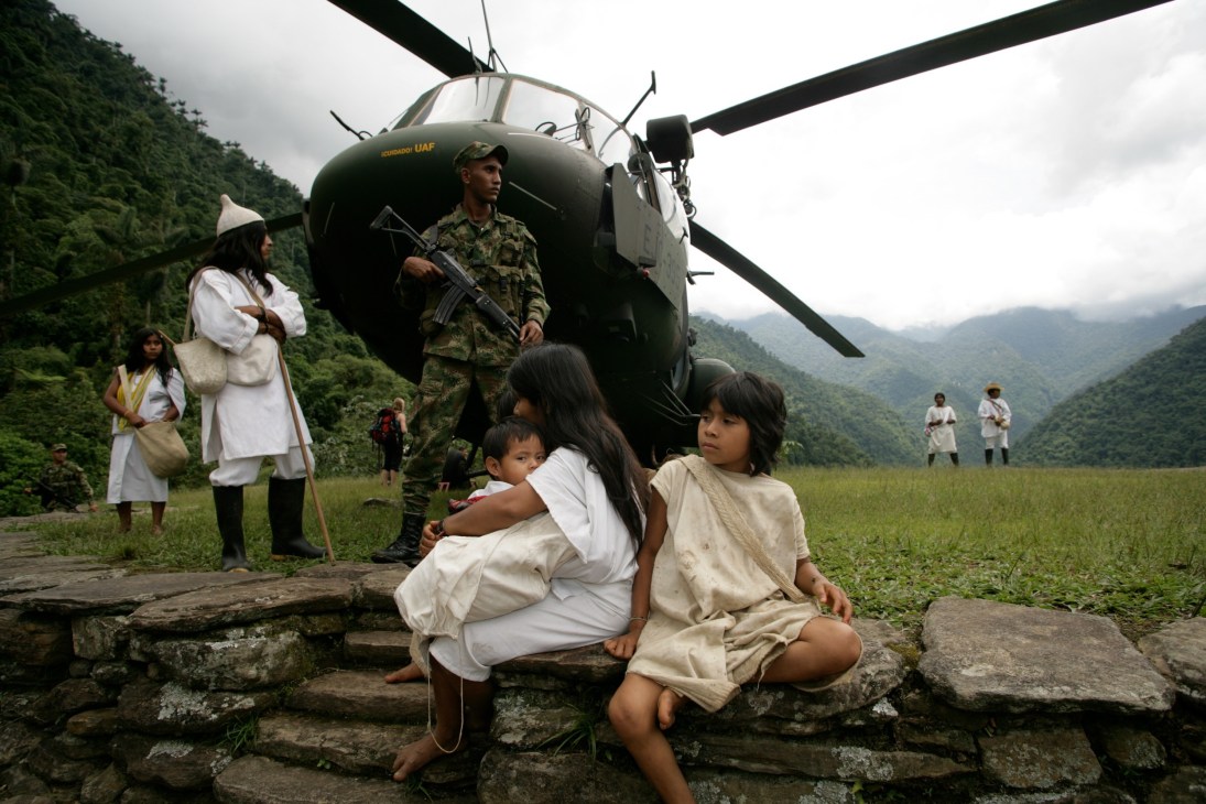 A armed soldier stands near a military helicopter as local civilians, including children in white clothing, sit on rocks in a mountainous jungle valley.