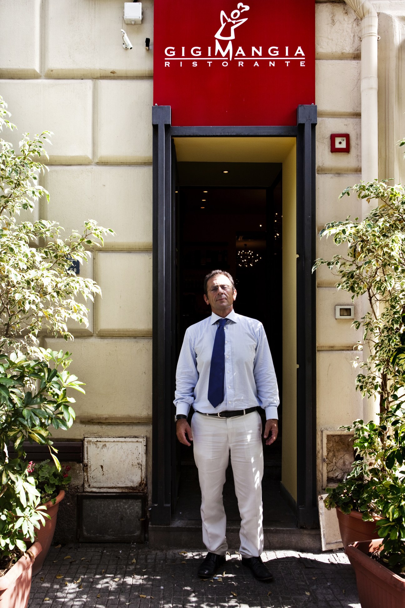 Gigi Mangia stands in the doorway of his Palermo restaurant beneath the red Gigi Mangia Ristorante sign.
