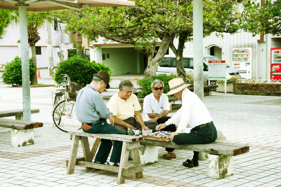 Four elderly men playing a game of Igo on a wooden bench in a shaded outdoor plaza in Naha.