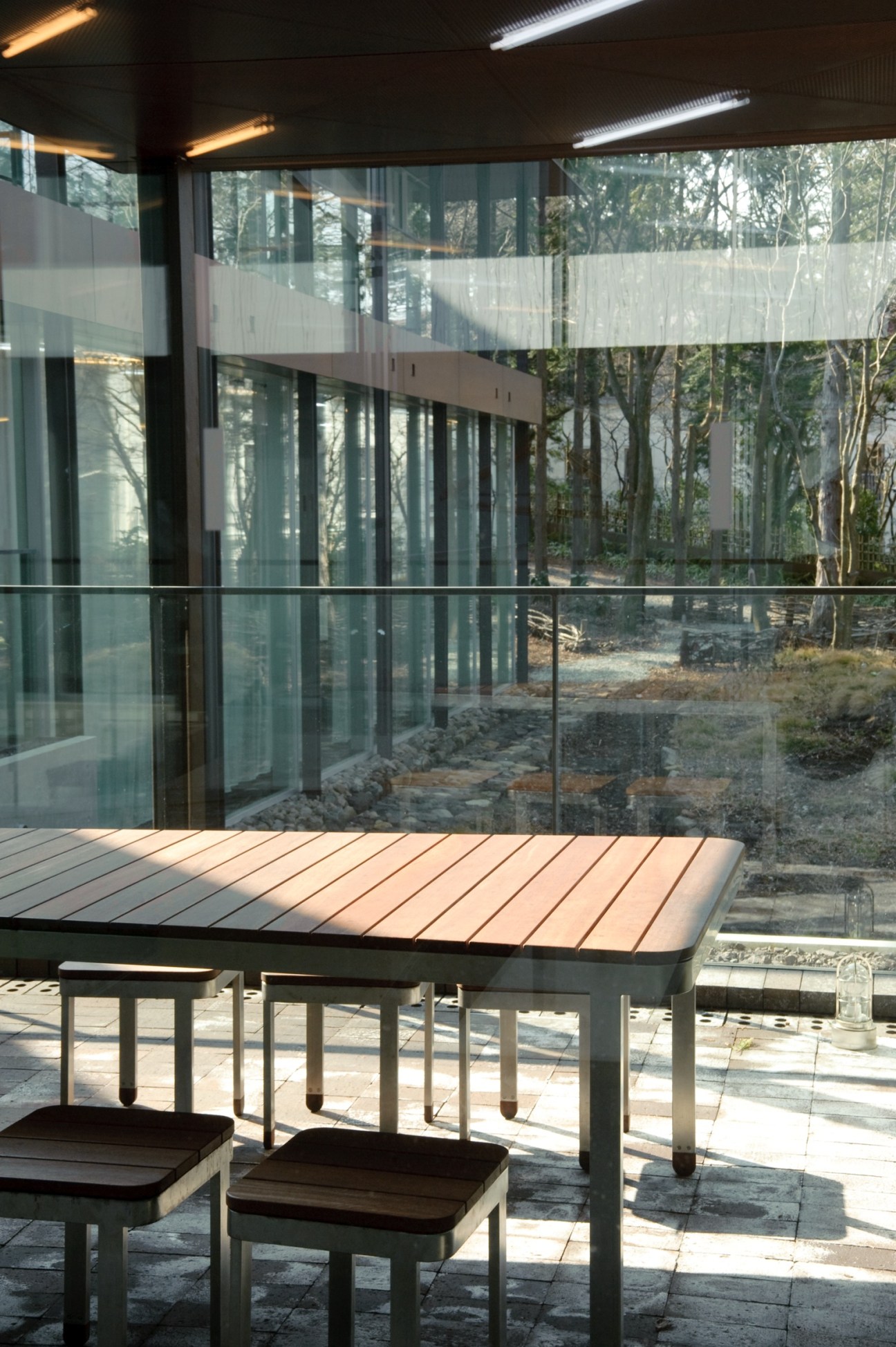 Modern wooden table and benches on a wooden deck with floor-to-ceiling glass walls overlooking a forested landscape.