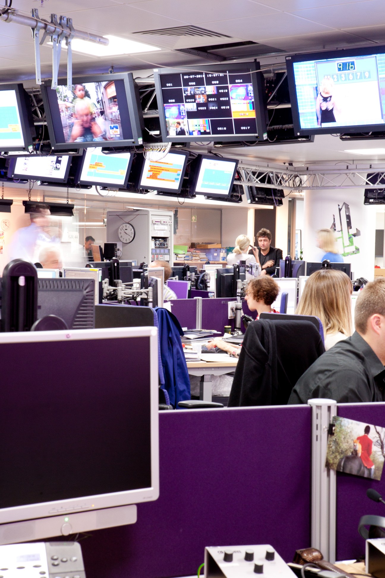 Channel 4 News newsroom with journalists at purple-partitioned desks and multiple monitors displaying content on the wall above.