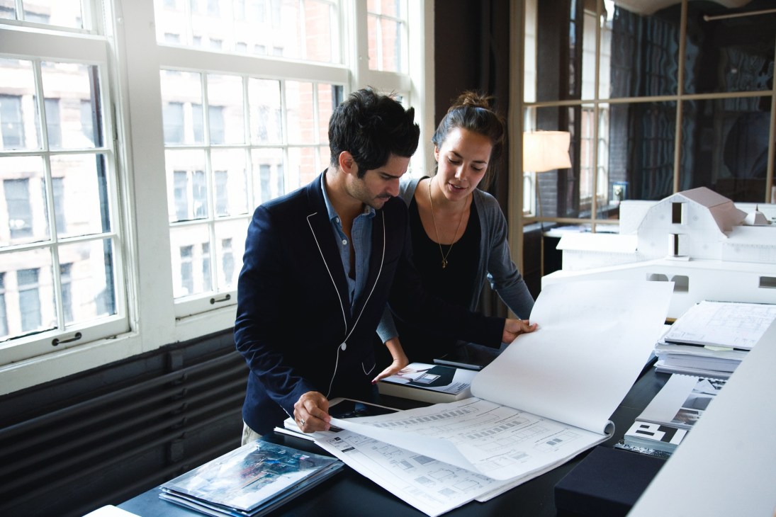 Two people review architectural plans and models at a desk in a bright office with large windows overlooking city buildings.