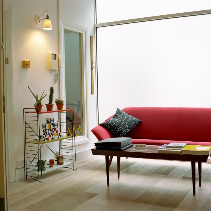 Modern reception area with red sofa, mid-century furniture, potted plants on wire shelf, and warm lighting.