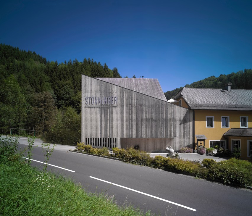Modern timber-clad distillery building with angled roof nestled in forested Austrian valley landscape.