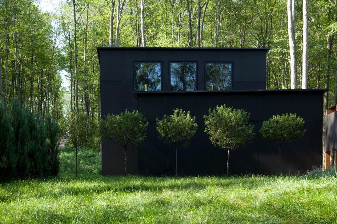 A black minimalist garage structure with three windows, surrounded by tall forest trees and landscaping shrubs.