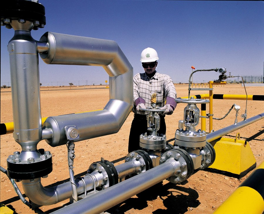 Worker in hard hat inspecting industrial piping and yellow equipment at a desert facility.