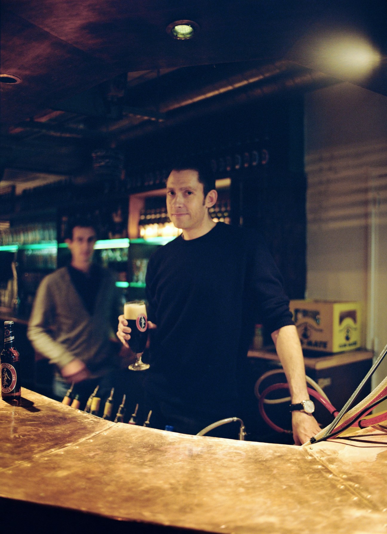 Two men at a bar counter, one holding a glass of dark beer, with bar equipment visible in background