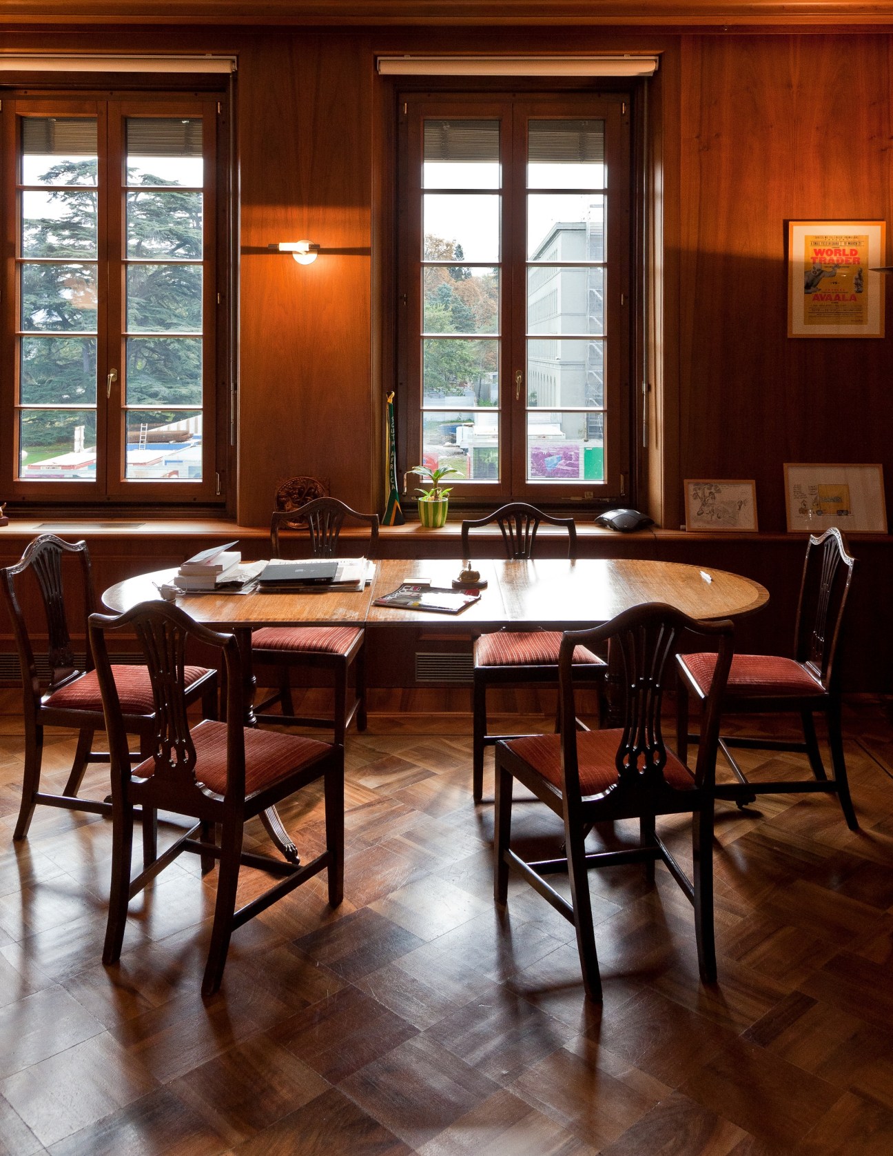 Wooden oval meeting table with chairs in a wood-paneled office with large windows.