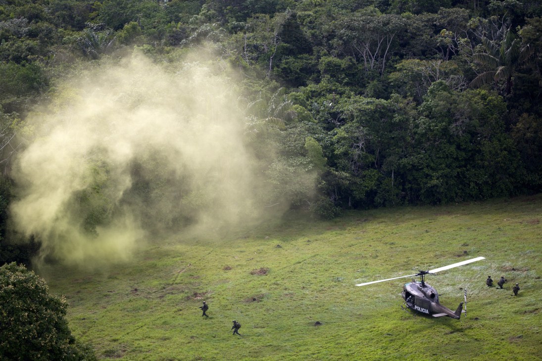 Colombian police helicopter landing in a jungle clearing with smoke from coca laboratory raid visible.