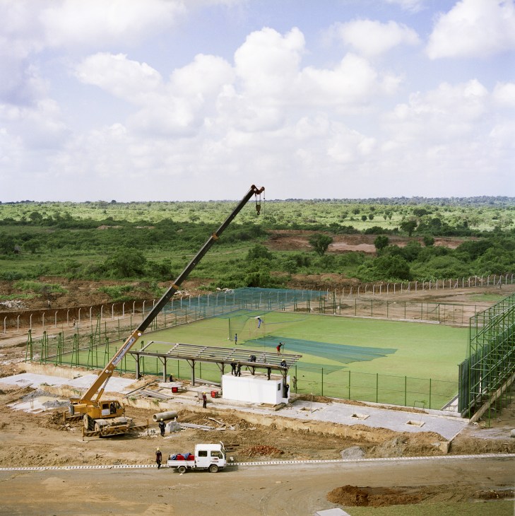 Cricket stadium under construction in rural Sri Lanka with crane, green field, and jungle landscape.