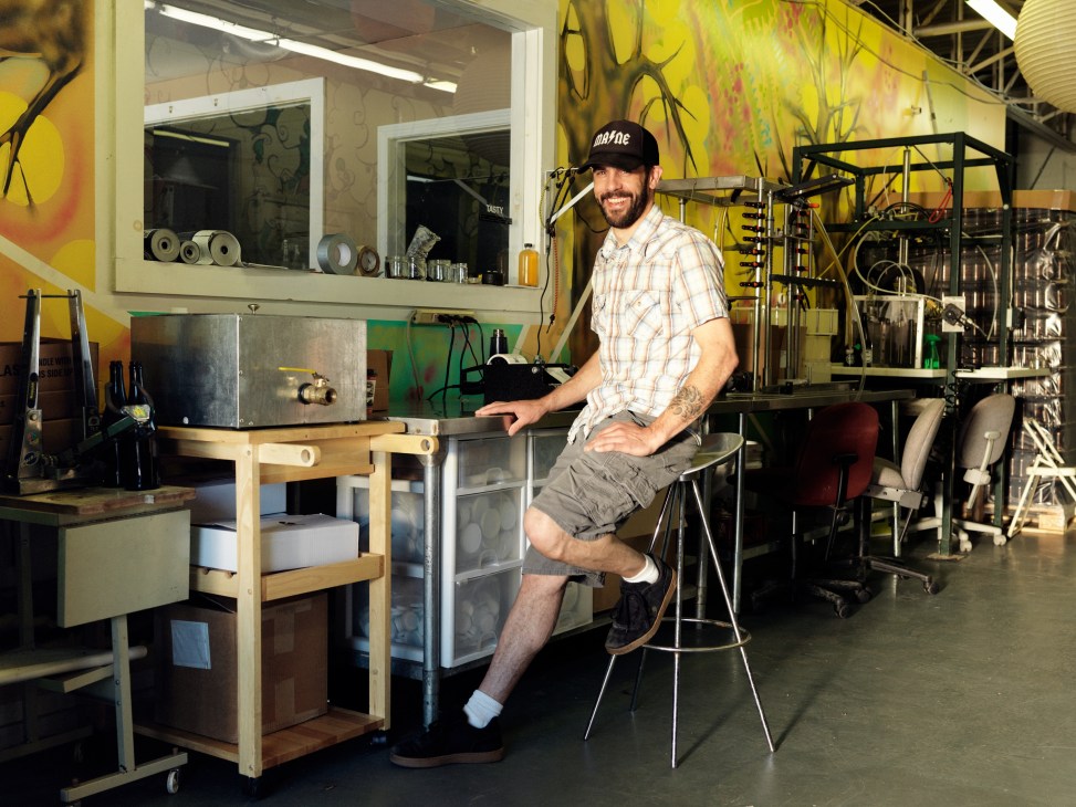 Eli Cayer sits on a stool at Urban Farm Fermentory, surrounded by fermentation equipment and psychedelic yellow wall art.
