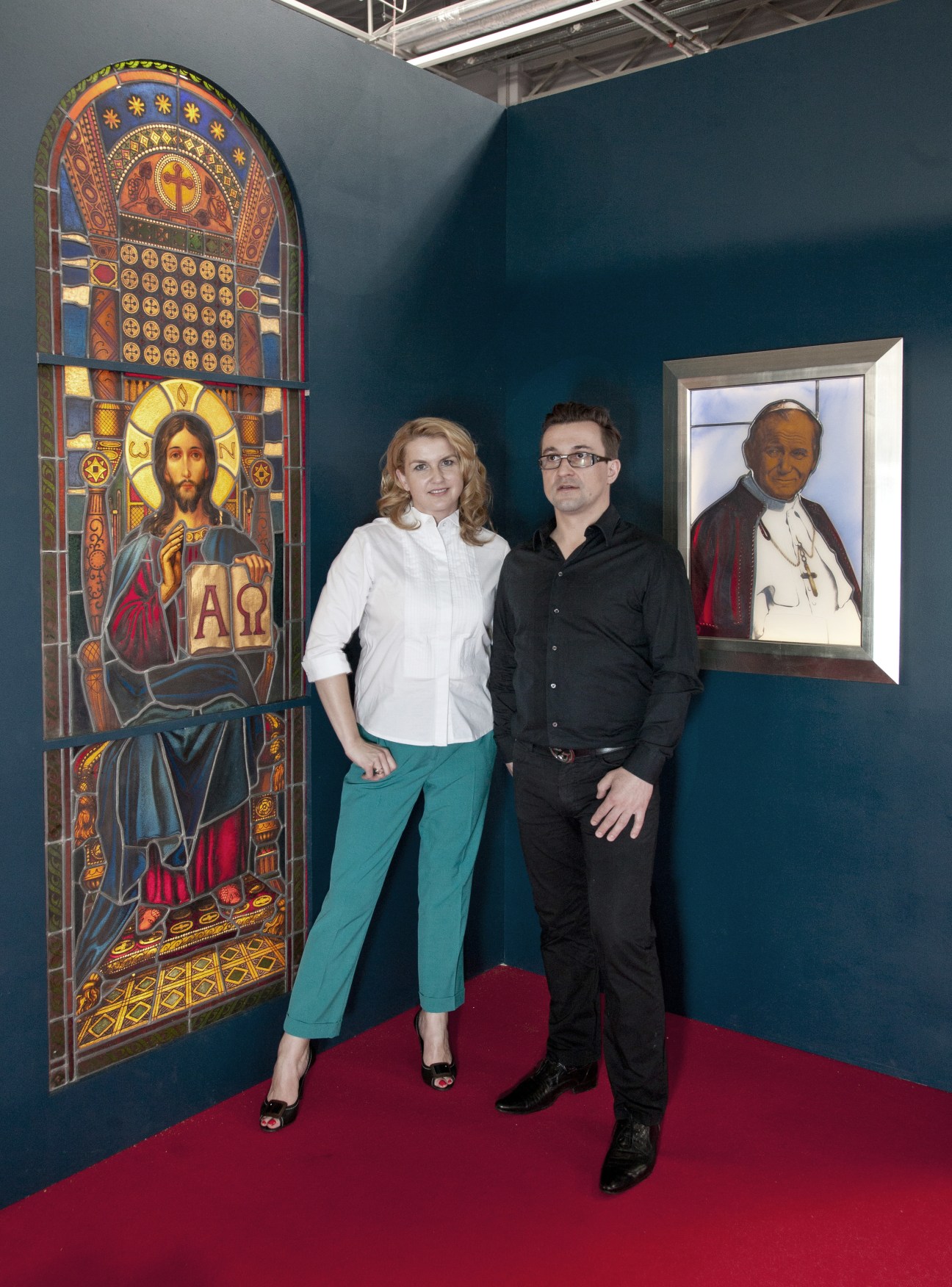 Piotr and Ursula Ostrowski stand with stained-glass windows including a Jesus Christ panel and Pope John Paul II portrait.