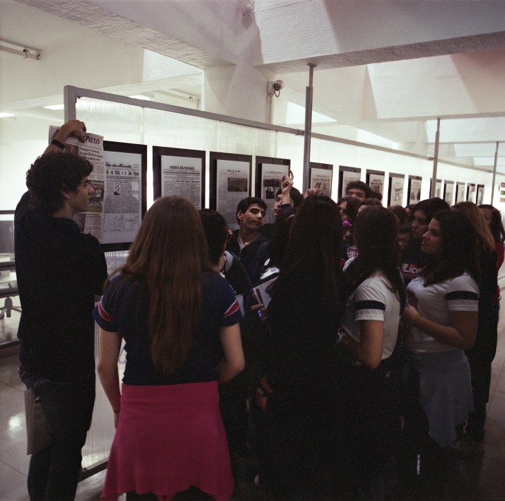 A group of students gathers in a bright newsroom viewing framed newspaper front pages on display walls.
