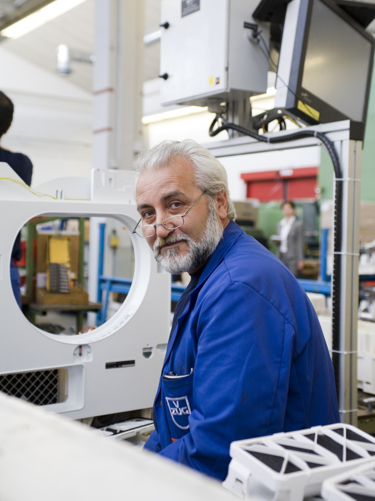 An older engineer with grey hair and glasses wearing a blue V-Zug jacket inspects white appliance components in the factory.