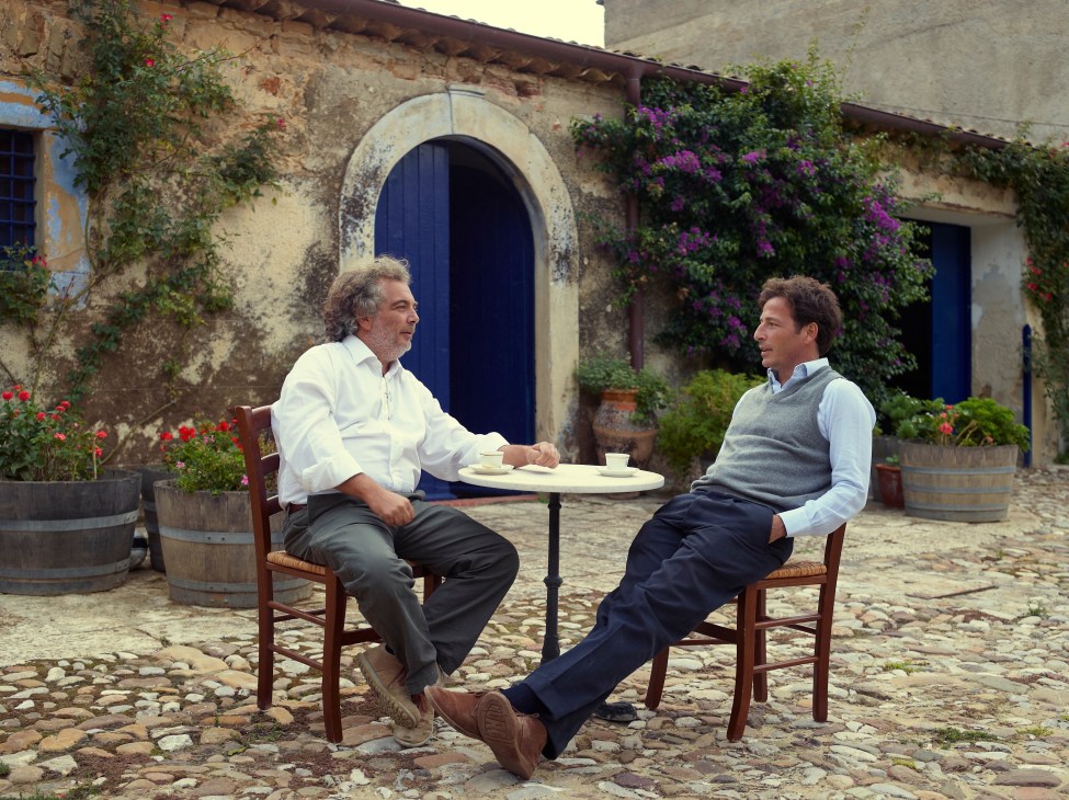 Two men seated at a small table in a courtyard of a historic stone building with purple flowers.