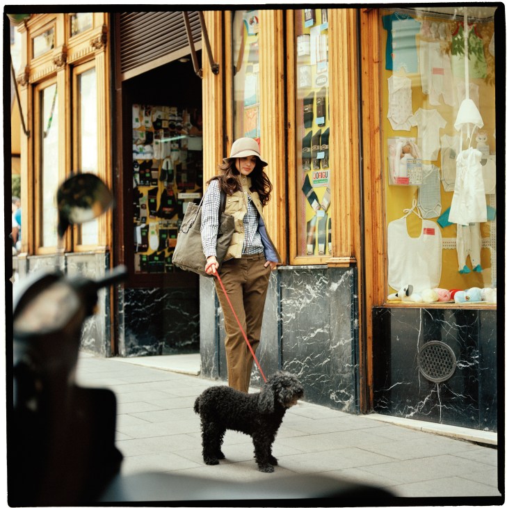 Woman walking dog past storefront wearing Toujours hat, Penfield vest, Melograno Editions shirt, C.P. Company trousers and Closed bag.