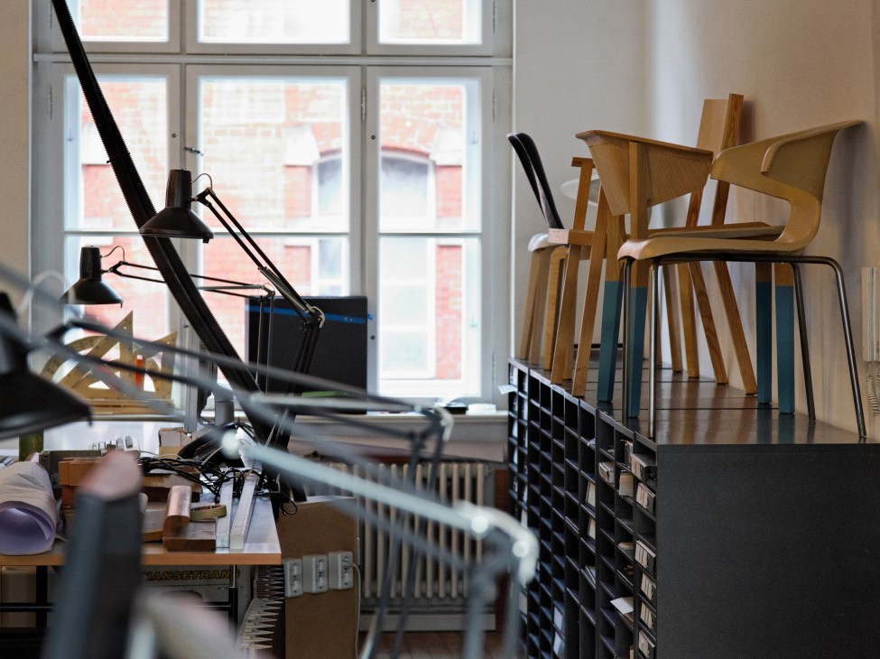 Wooden chair prototypes displayed by a window in a design studio with desk lamp and materials.