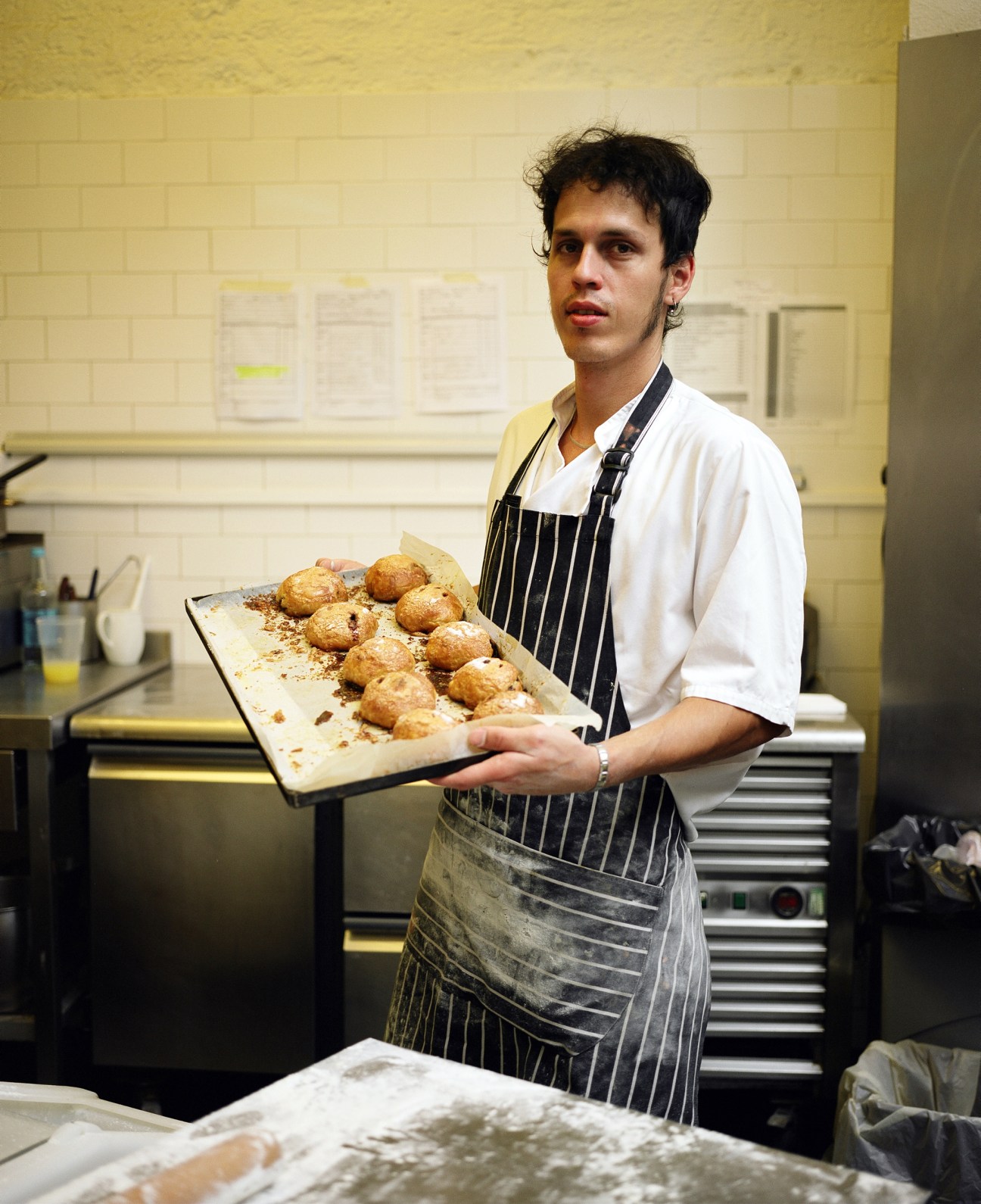 Pastry chef Fabio Ferreira Carvalho holds a tray of baked pastries in a kitchen.