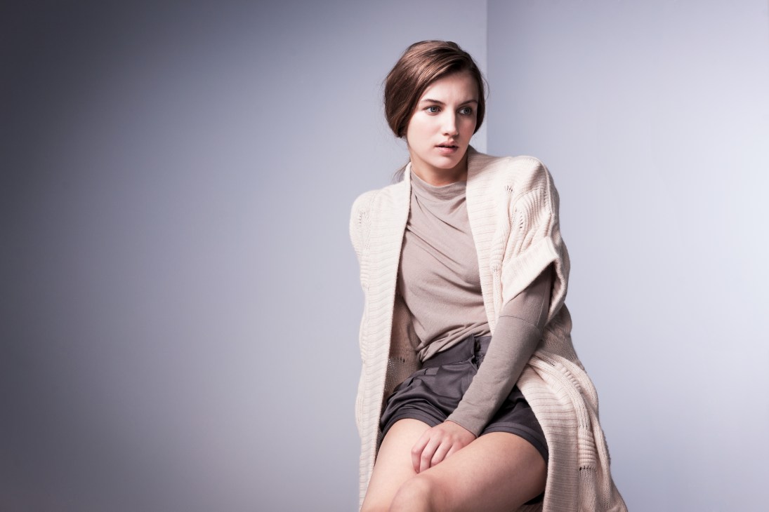 Woman wearing a cream-colored cardigan over a taupe turtleneck and dark shorts against a neutral background.