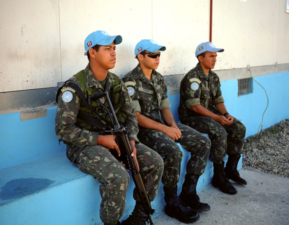 Three Brazilian UN peacekeeping troops sitting on a bench wearing camouflage uniforms and blue UN caps.