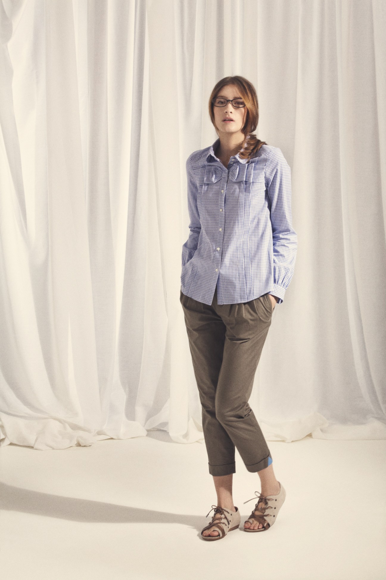 Woman wearing purple gingham shirt and olive chinos standing against white curtain backdrop.