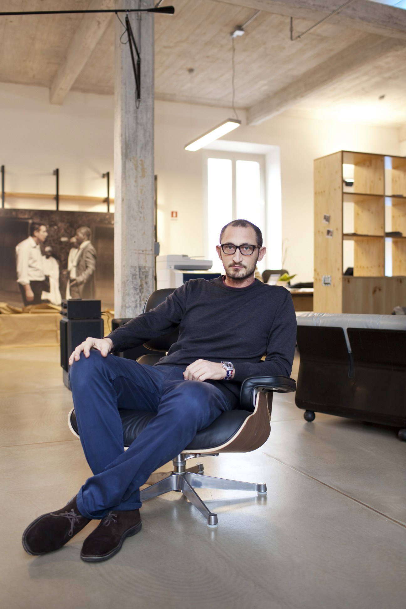 Man in glasses sitting in modern office with exposed concrete beam and wooden shelving.