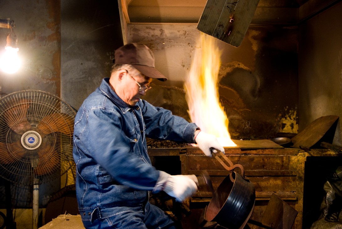 Lee Bong-ju working with glowing metal in his bronze workshop, shaping a piece over a forge.