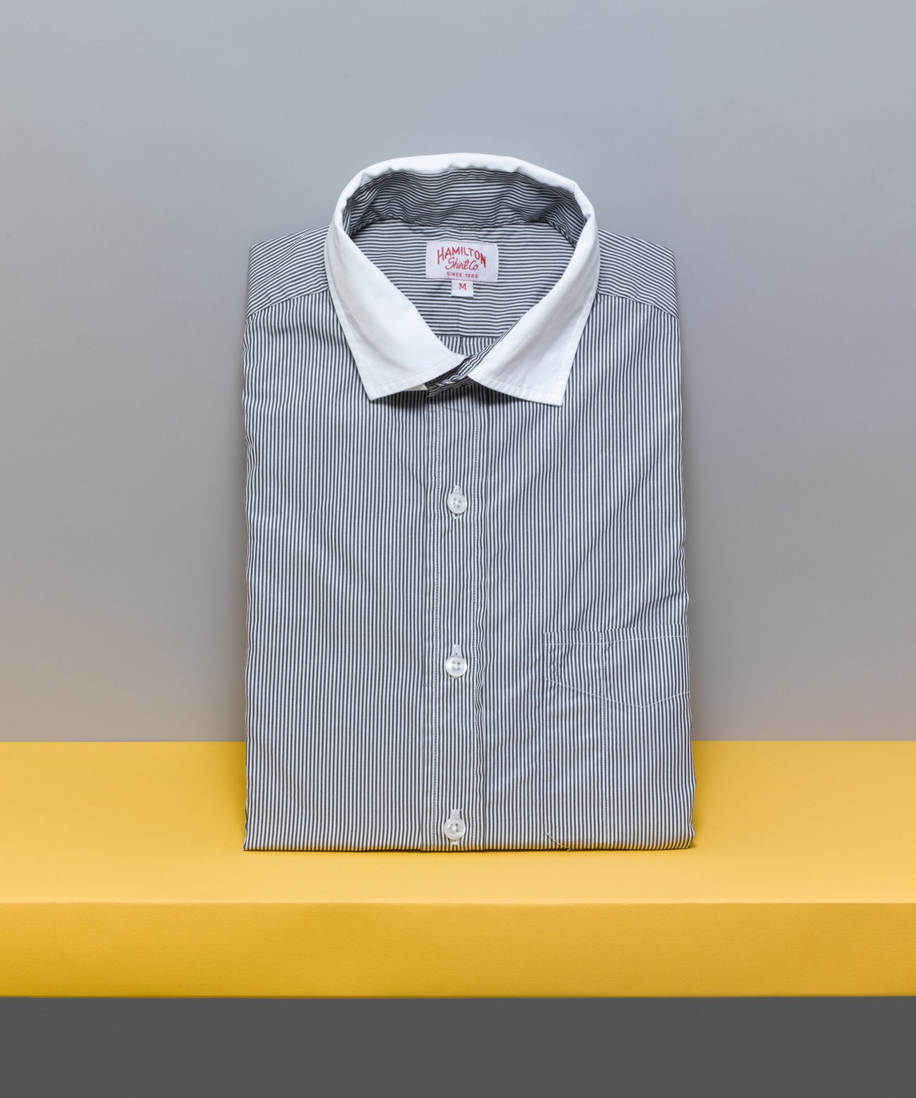 Folded Hamilton 1883 striped shirt with white collar displayed on yellow and grey background.