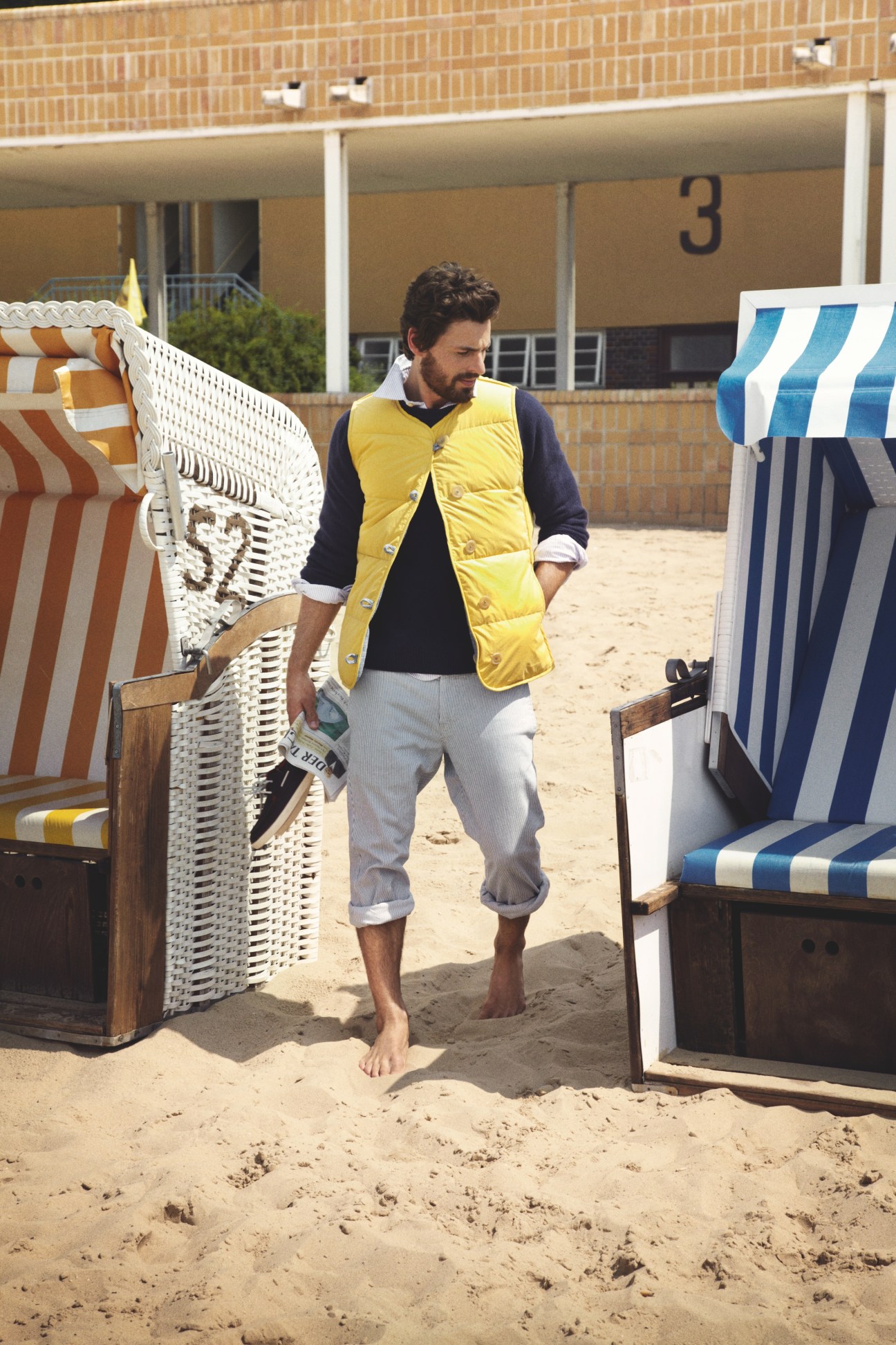 Man wearing yellow puffer vest and light trousers standing on beach near striped beach chairs.