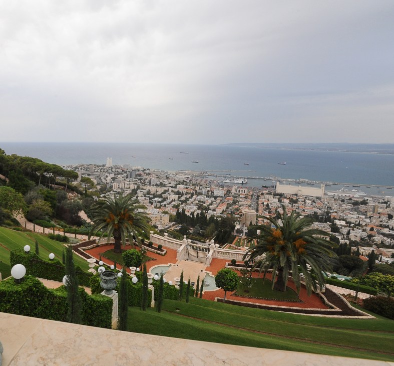 Aerial view of Haifa cityscape with port, Mediterranean Sea, and Carmel mountains with palm trees in foreground.