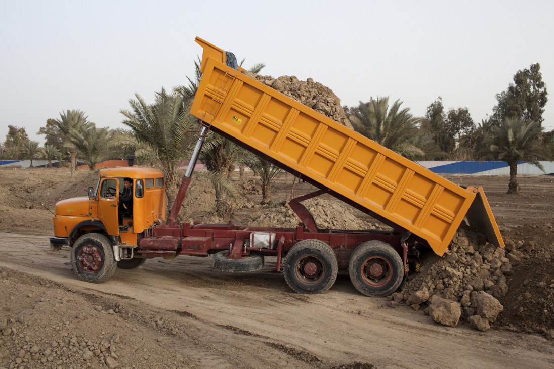 Yellow dump truck unloading soil at a Baghdad beautification project surrounded by palm trees.