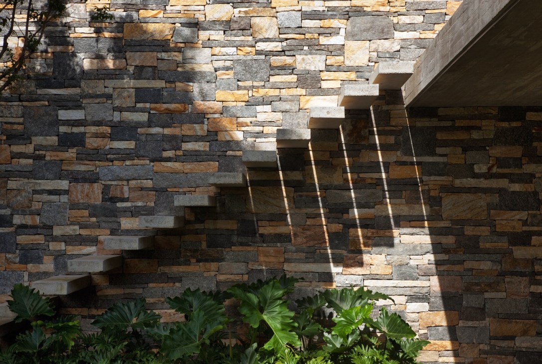 Stone wall with stairway and green plants, shadows cast by sunlight through overhead structure.