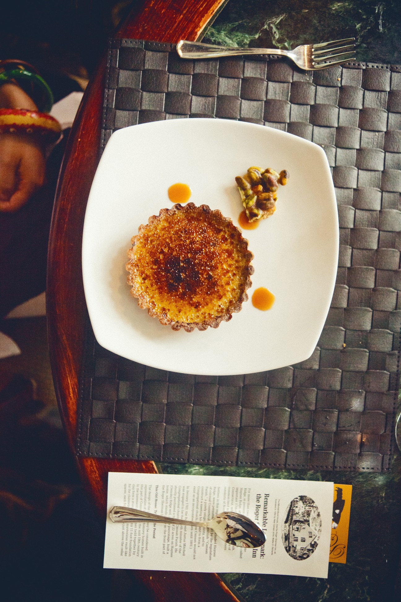White chocolate and pistachio crème brûlée dessert on a white square plate with decorative orange dots and pistachio garnish.