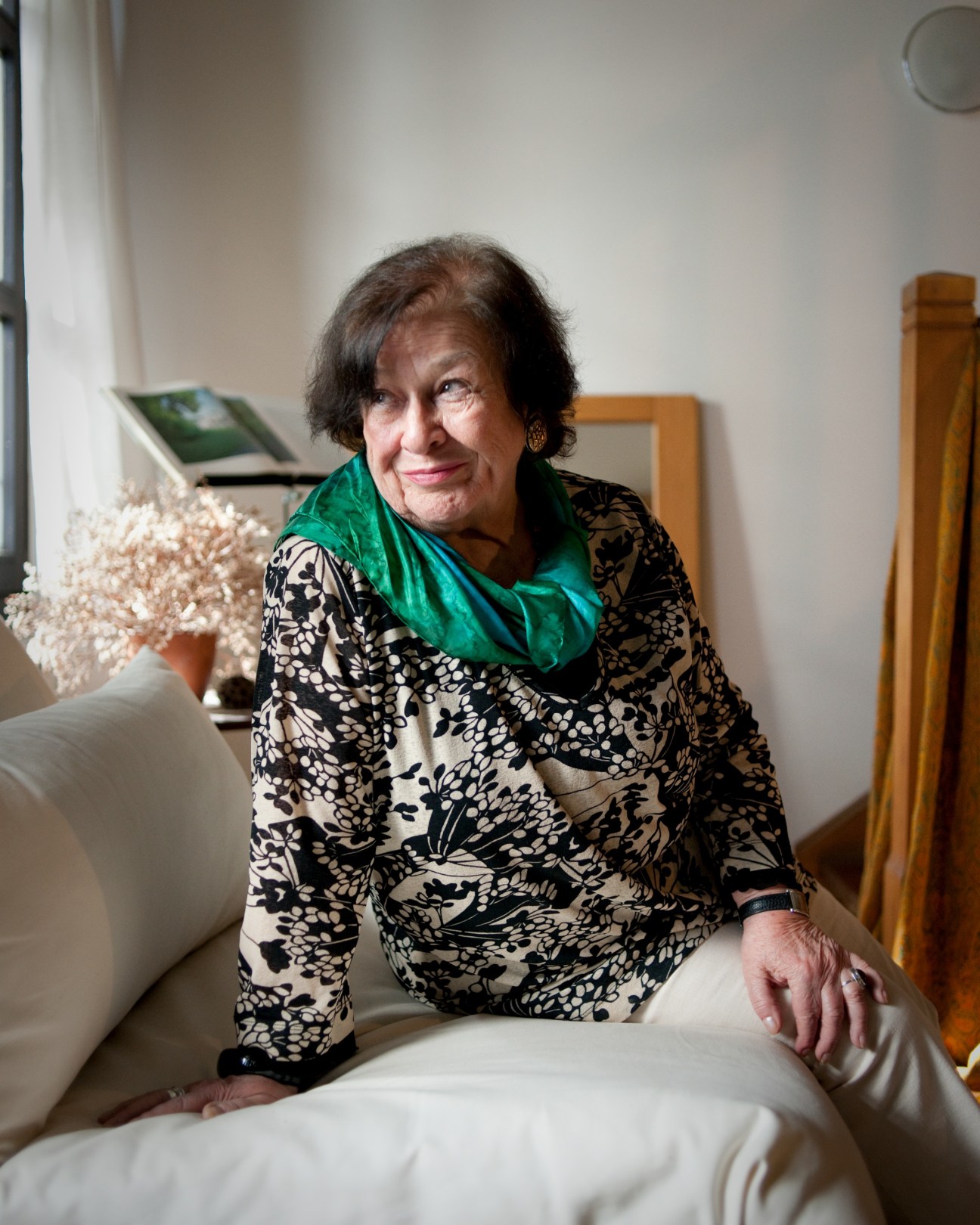 Elderly woman in patterned black and white jacket with green scarf sitting indoors