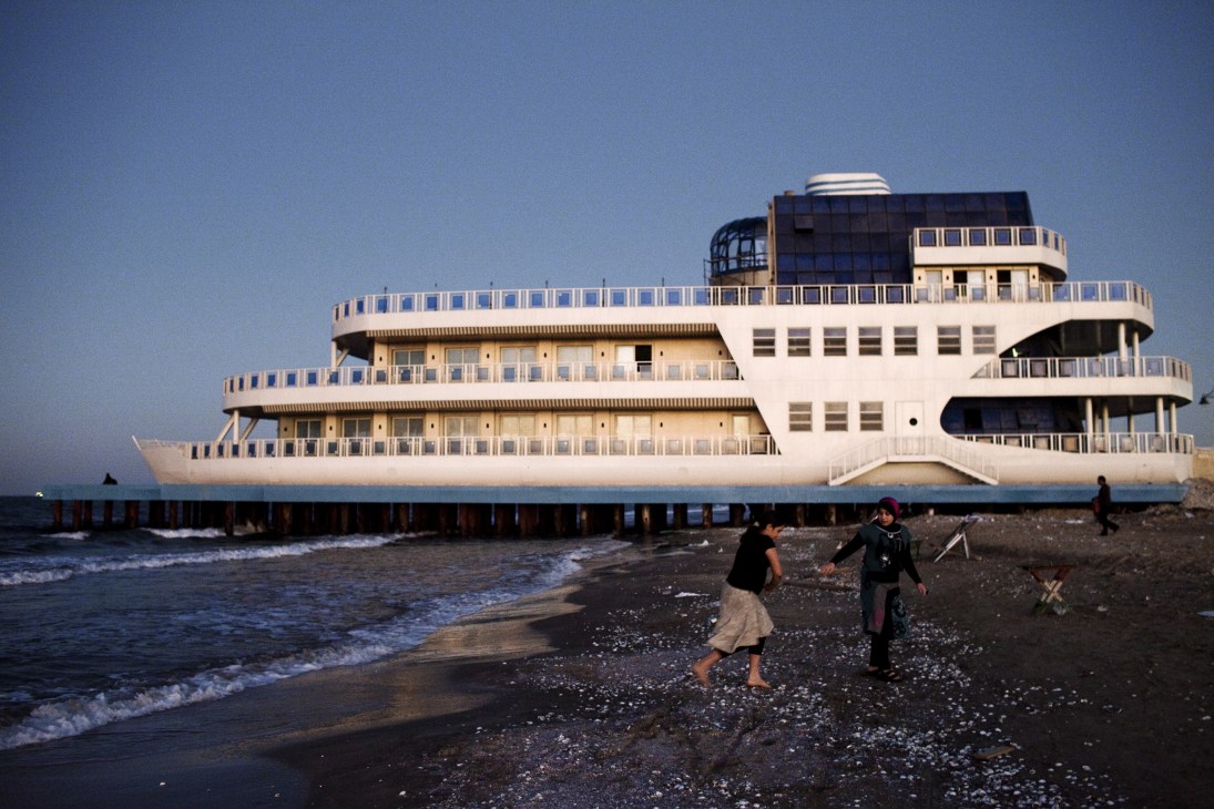 A large multi-story ship-shaped structure under construction on a beach in Port Said, with two people walking on the shore.