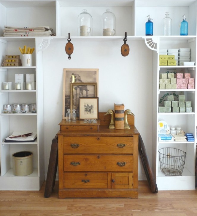 White shelving display with vintage wood dresser featuring curated products including soaps, bottles, textiles and home goods.