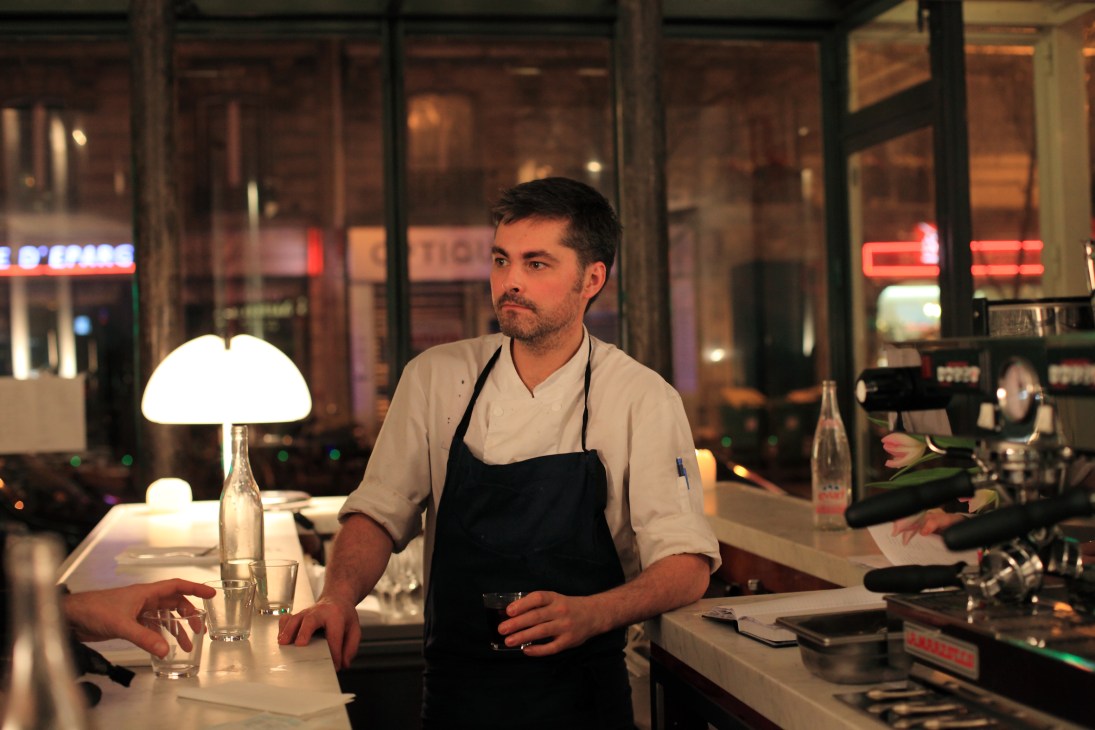 Chef at Le Dauphin restaurant standing behind white marble bar at night with city lights visible through windows.